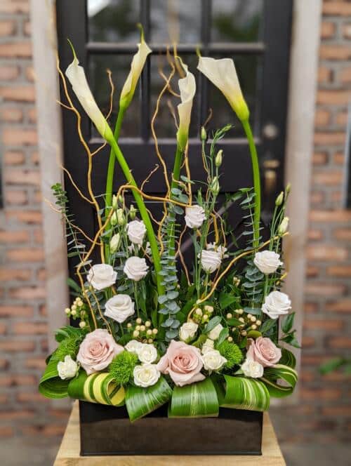 The Watering Can | Tall white callas surrounded by pink and white florals and textured greens in a black rectangular tin.