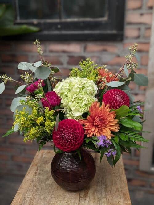 The Watering Can | A colourful bouquet of hydrangea, dahlia, mums, and garden greens in a round burgundy vase.