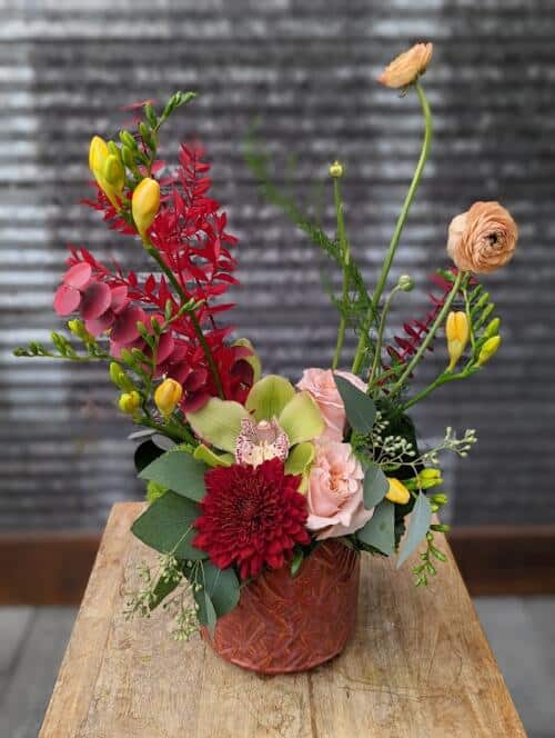 The Watering Can | A red, peach and yellow floral arrangement in a dark orange container.