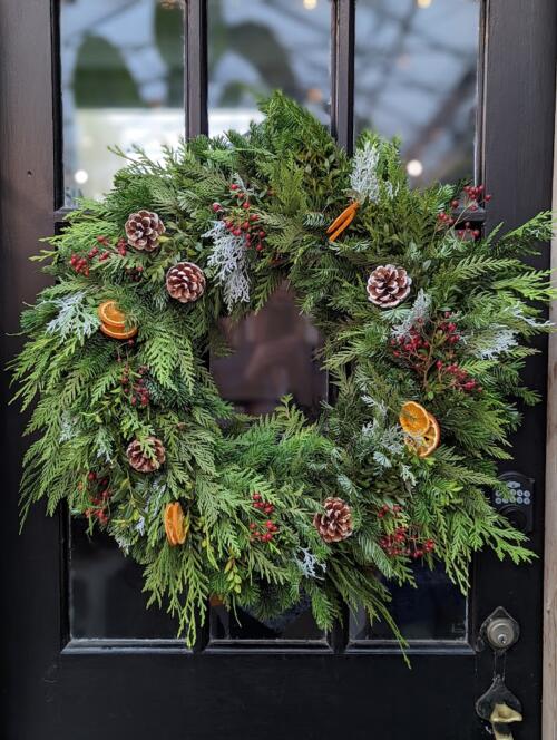 The Watering Can | A mostly cedar wreath with flecks of white abvardia and boxwood, decorated all around with pine cones, orange slices, and rosehips.
