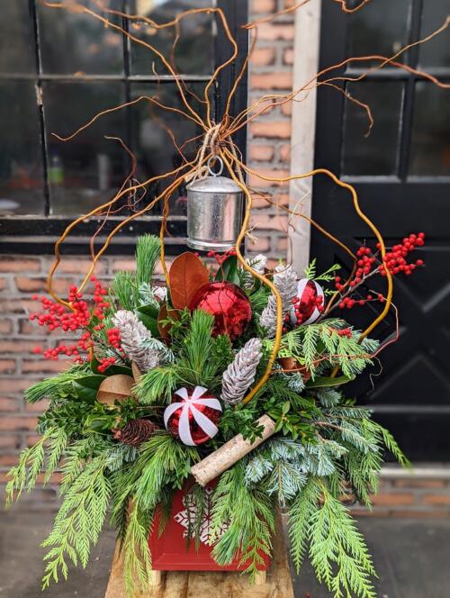 The Watering Can | A red and white winter urn design featuring a large silver bell designed in a decorative square red container.