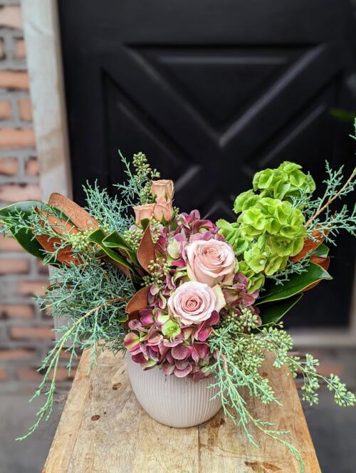 The Watering Can | A bouquet in vase made with winter greens, magnolia, hydrangea, and roses, in blush tones designed in a round white ceramic vase.