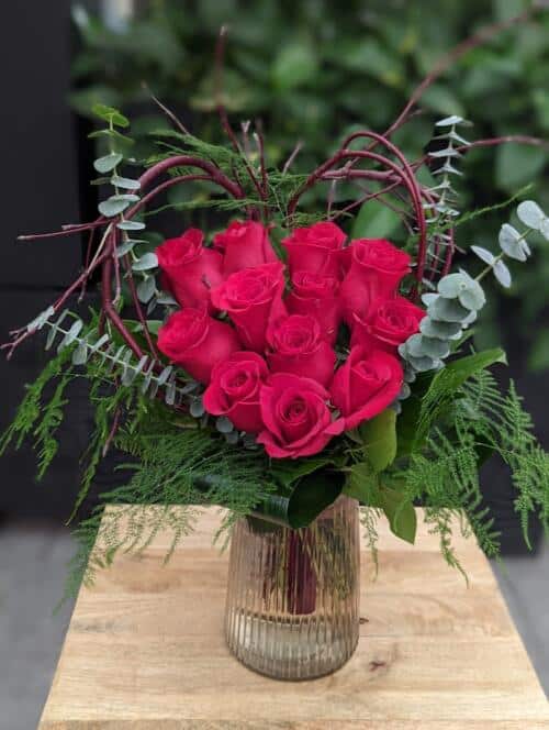 The Watering Can | A dozen red roses arranged in the shape of a heart surrounded by a red dogwood heart and greens in a glass vase.