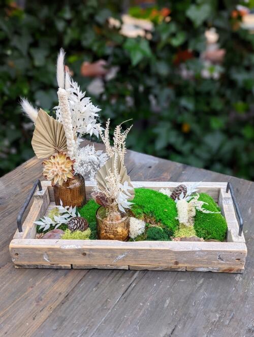 The Watering Can | A wooden tray with 2 small brown bud vases containing natural dried items surrounded by moss and stones.