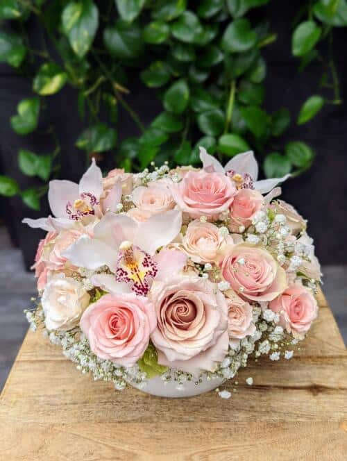 a white bowl filled with pink and white blooms, including roses, spray roses, orchids, and hydrangeas