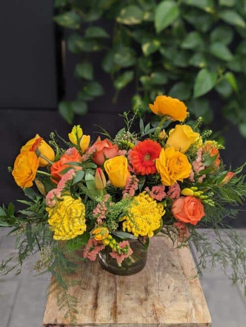 The Watering Can | A flowing garden bouquet in orange, and yellow in glass cylinder vase.