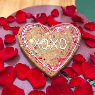 A heart-shaped cookie decorated with pink XOXO icing, surrounded by red rose petals on a pink plate.