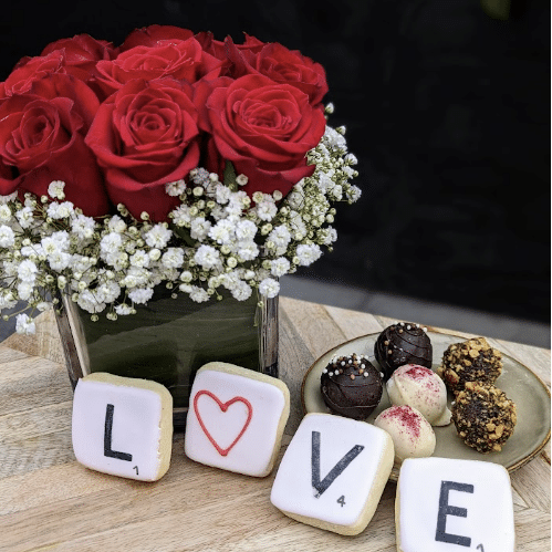 A romantic Valentine's Day arrangement of red roses and baby's breath beside Scrabble tiles spelling LOVE and chocolate truffles on a wooden surface.