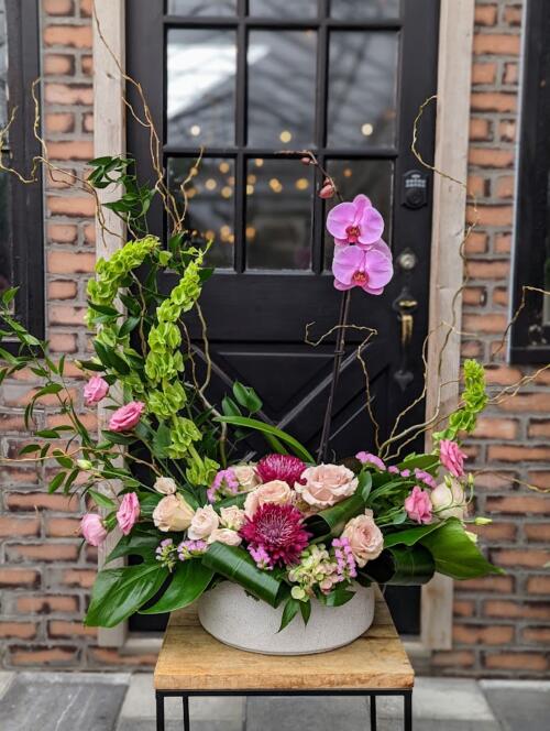 A large round grey ceramic container filled with purple mums, pink lisianthus, soft pink roses and spray roses. featuring tall green bells of ireland, leafy italian ruscus, spiraling curly willow branches and a bold pink orchid.