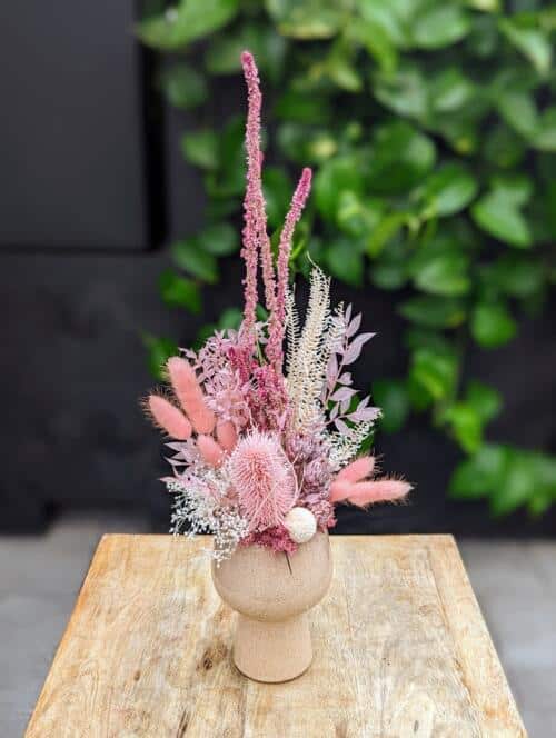 A beige pedestal container filled with pink dried blooms. A blend of textures keeping this arrangement unique from every angle.