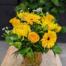 A yellow prisim glass vase with a silver handle. Bursting with yellow blooms of roses, gerberas, freesia, chamomile and mums. Regular greenery and bupleurum.