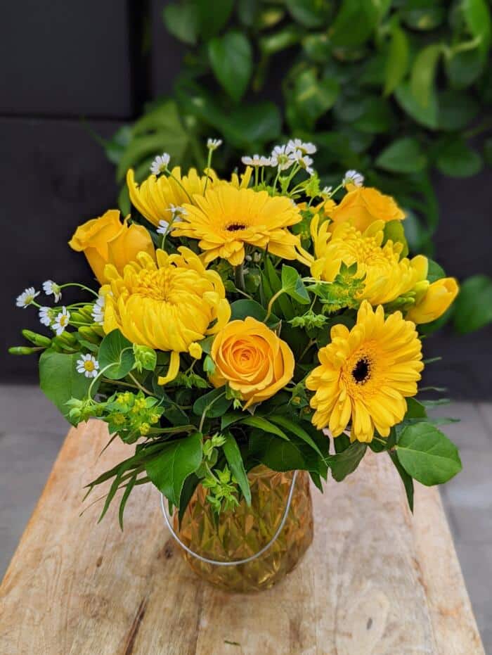 A yellow prisim glass vase with a silver handle. Bursting with yellow blooms of roses, gerberas, freesia, chamomile and mums. Regular greenery and bupleurum.