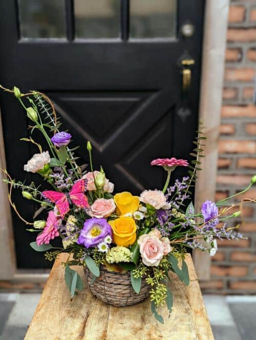 A grey woven basket bursting with asymmetrical blooms. Featuring bright yellow roses, soft pink spray roses, purple lisanthus, baby blue eucalyptus, seeded eucalyptus and more. A very spring colour palette.