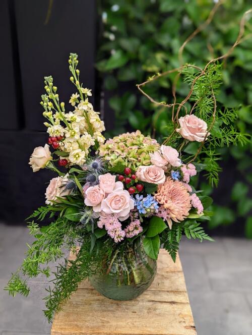 An asymmetrical bouquet in vase overflowing with woodsy flora. Featuring mojito hydrangea, dusty roses, spray roses and a mirage of greens to add whimsy. A blend of greens, blush and hints of purple thistle and burgundy.