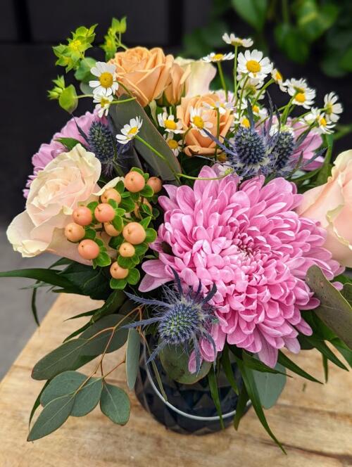 A spring bouquet in a blue glass vase. Featring lavender mums, light pink roses, thistle, chamomile, peach hypericum and delicate chamomile.