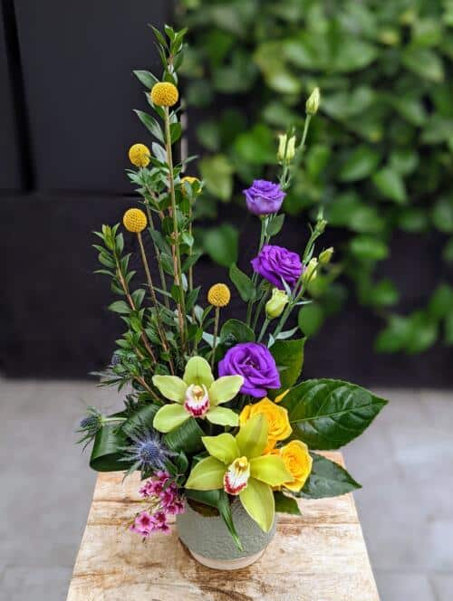 A blue/green textured container with reaching purple lisianthus, yellow craspedia, green orchids, and yellow roses.