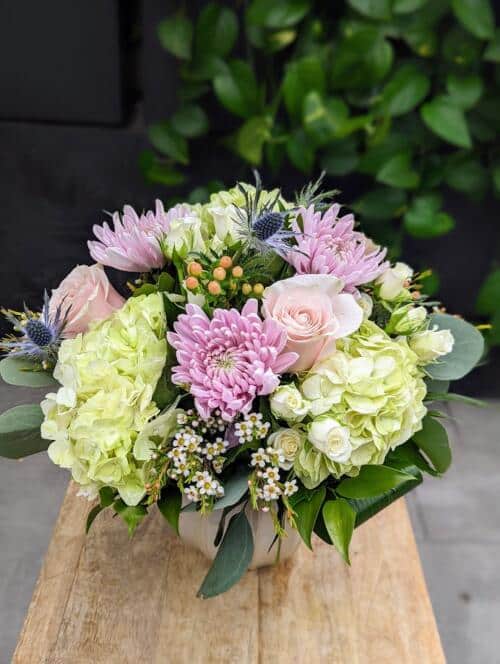 A purple and green bouquet in a low taupe ceramic container. Featuring green hydrangeas, light purple mums, light pink roses, peach hypericum and more.