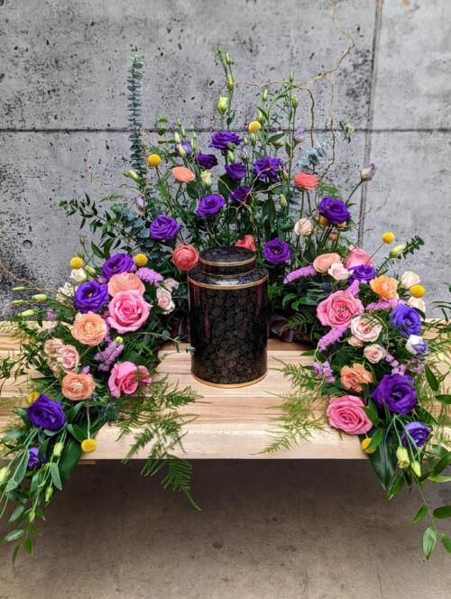 A cremation arrangement of wrapping pruple lisanthus, pink roses, peach ranunculus, yellow craspedia, thist and an abundance of greenery.