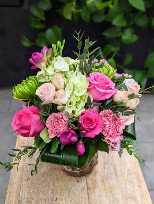 A light pink glass vase filled with green and pink blooms such as green hydrangea, hot pink roses and so much more to brighten your day.