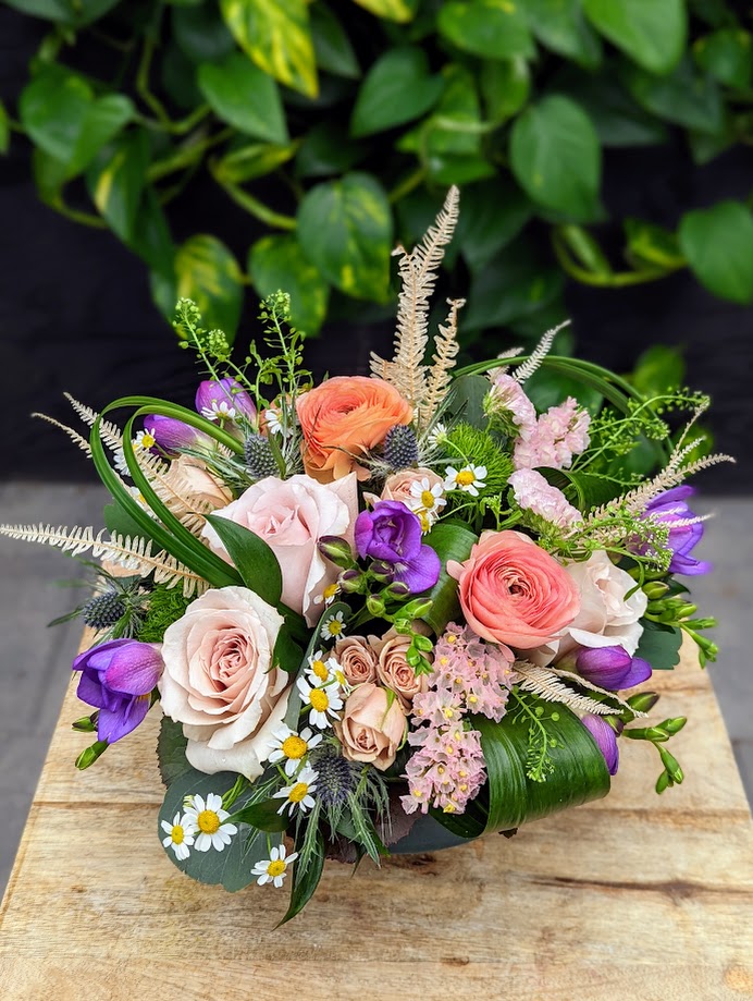 A soft pastel european arrangement overflowing with blooms. Featuring peach ranunculus, purple lisianthus, quicksand roses, chocolate spray roses, purple fragrant freesia, and many more blooms. Reminiscent of a fairy tale.