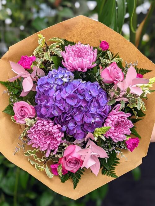 A purple and pink bouquet. Featuring a dark purple hydrangea, light purple mums, light pink israeli ruscus, pink roses and variegated greens.