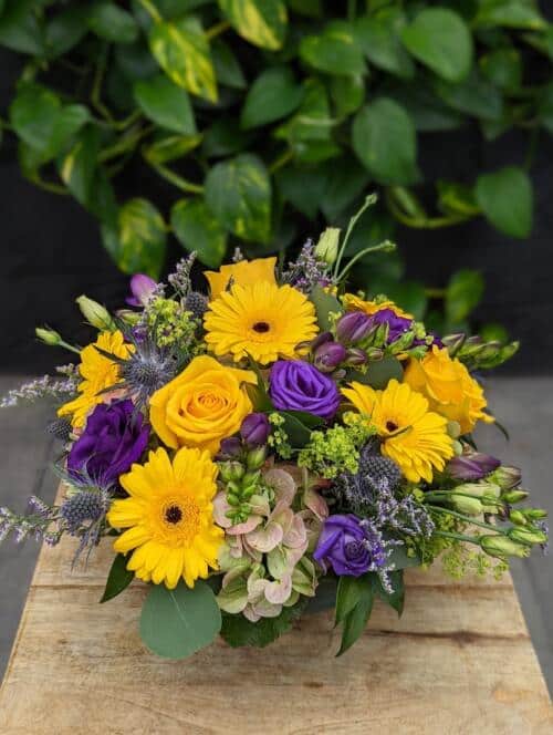 A large blue ceramic bowl filled with yellow and purple blooms. featuring bright yellow roses, yellow mini gerberas, deep purple lisianthus, fragrant purple freesia and sprigs of purple limonium.