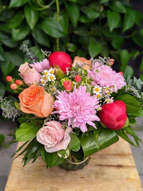 a light blue vase with handle, lavender mums, a mix of pink and peach roses, red peonies, purple limonium, peach hypericum and more.