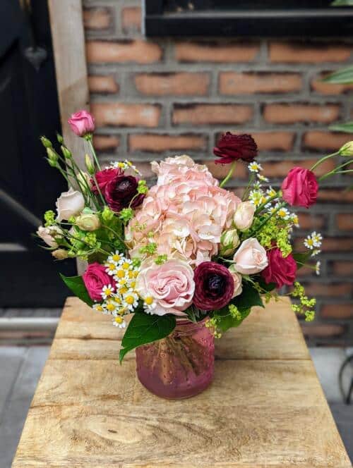 A petit pink glass jar filled with pink and burgundy blooms. featuring brugundy ranunculus, dark pink lisianthus, pink hydrangea, dainty chamomile and more..
