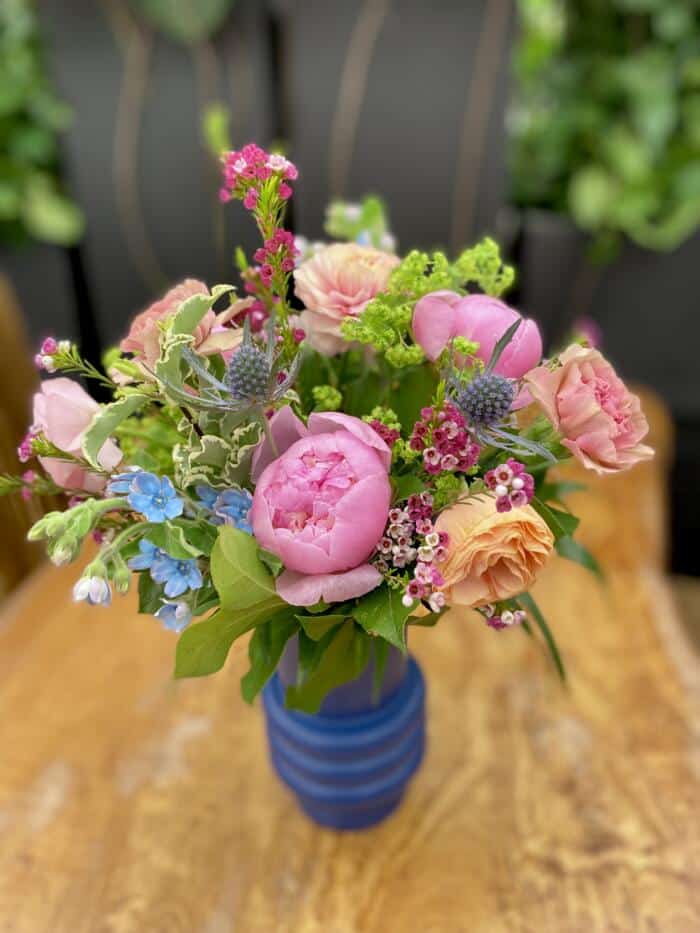A blue ringed glass vase filled with pink and peach blooms and a variety of greenery. featuring large pink peonies, peach ranunculus, dusty rose carnations, blue tweedia, purple thistle and more.