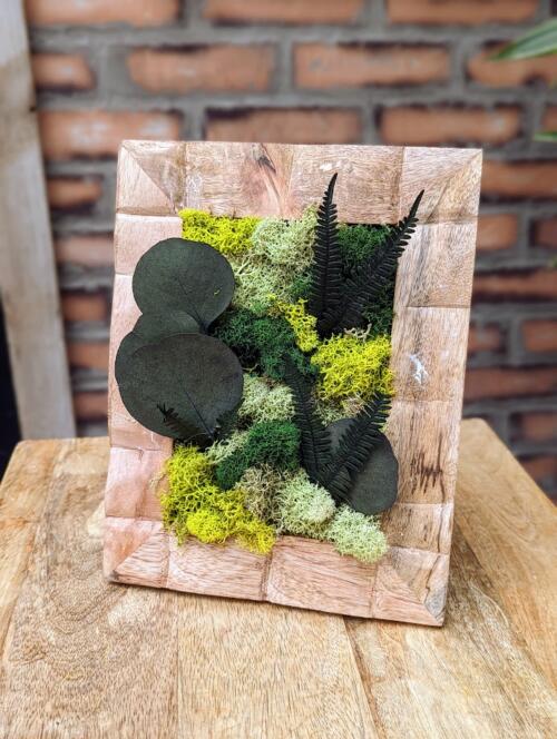 a light wood frame featuring a blend of green mosses and eucalyptus.