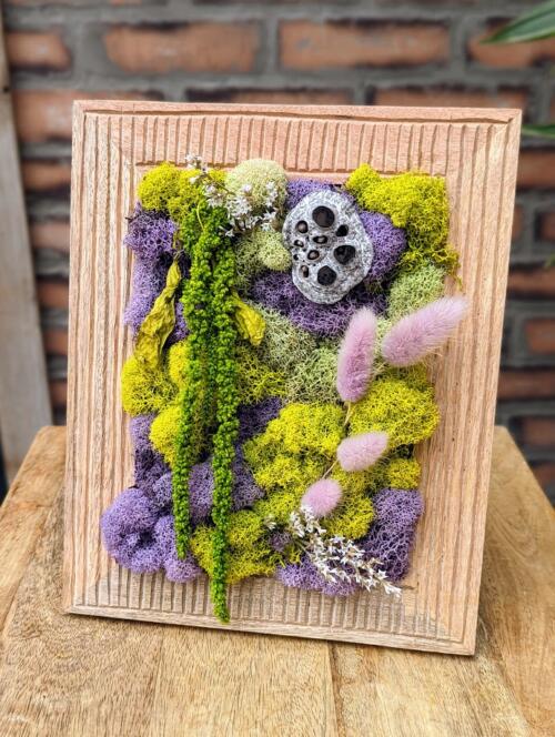 a light wood frame with lime green and lavender purple mosses adorned with a lotus pod and hanging amaranthus.