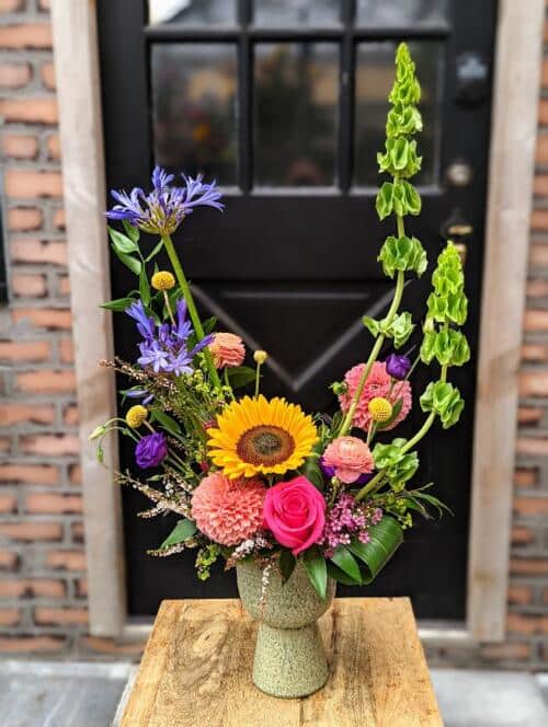 A green pedestal ceramic filled with reaching flower sof bellas of ireland, agapanthus, bright sunflowers. a mix of peach ranunculus, hot pink roses and pink dahlias.