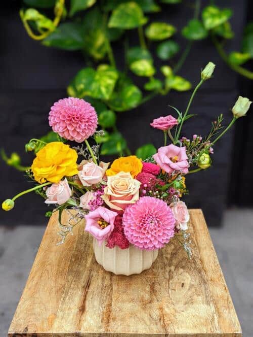 A beige ceramic container filled with bright yellow ranunculus, pink dahlias, pink lisianthus, light pink spray roses and a collection of roses.