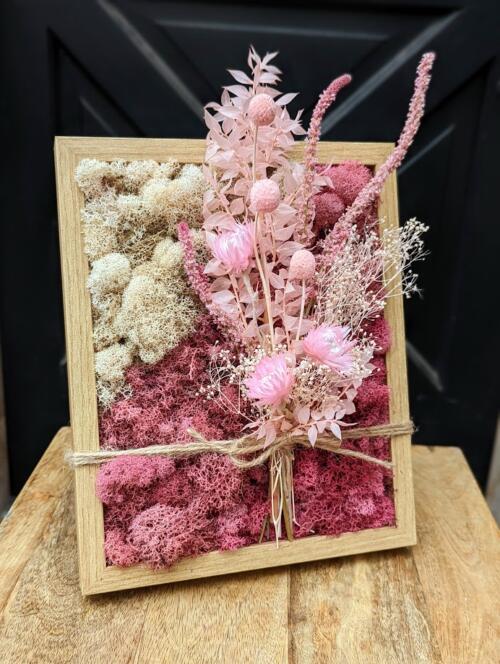 a light wood frame filled with sahdes of pink and white moss tied with a pink dried arrangement.