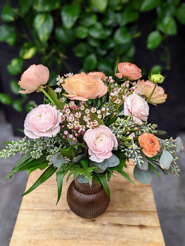 a brown ribbed vase filled with white, pinka dn peach ranunculus with tiny white waxflowers and seeded eucalyptus