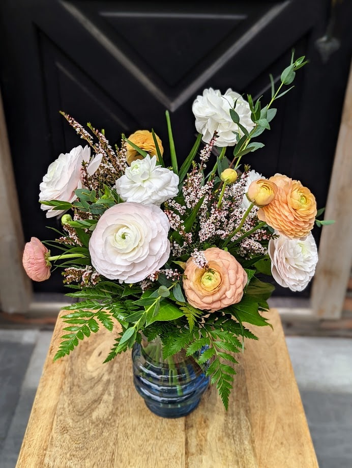 A blue glass vase filled with peach, white and blush ranunculus adorned with thryptomene.