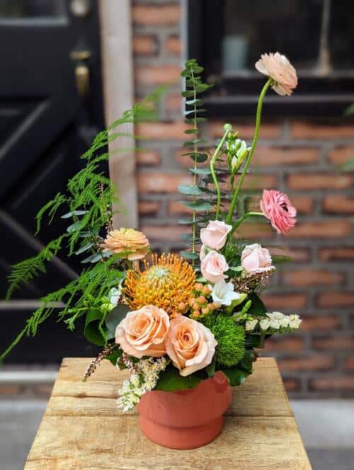 A coral ceramic filled with orange and peach blooms. featuring shimmer roses, blush spray roses, orange pin cushion and more