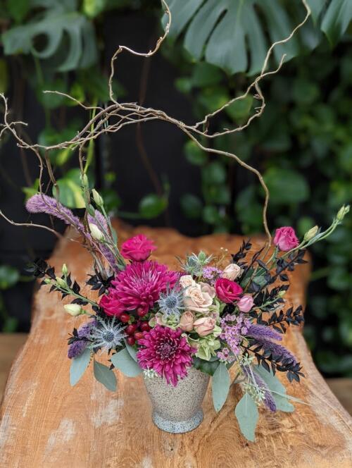 A light grey ceramic container overflowing with dark purple and pink blooms featuring mums, roses, waxflower and a mix of dried goods