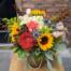 An amber vase with a rope handle overflowing with fall toned blooms. featuring rust coloured mums, yellow sunflowers, thistle, peach carnations, brown eucalyptus and a large antique hydrangea.