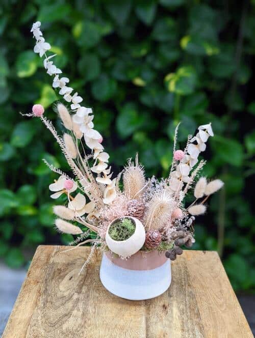 a cream and white ceramic container filledw ith light pink and cream dried florals in an asymmetrical design.