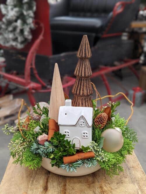 A neutral ceramic bowl filled with winter greens adorned with glass trees and a white ceramic house. adorned with a variety of ornaments