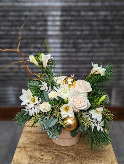 a beige container filled with winter greens and white florals adorned with gold ornaments.