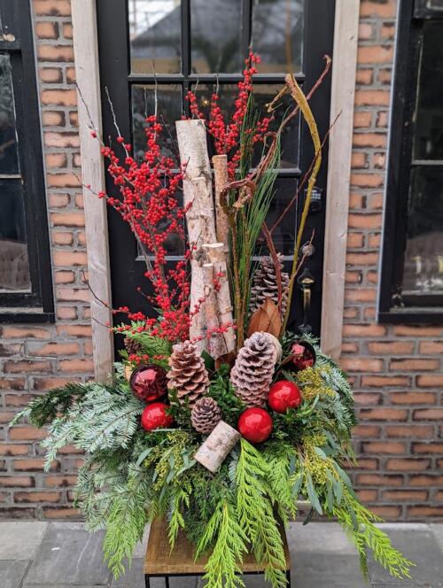 a large urn insert overflowing with winter greenery, reaching birch poles and ilex berries adorned with pinecones and assorted red ornaments.