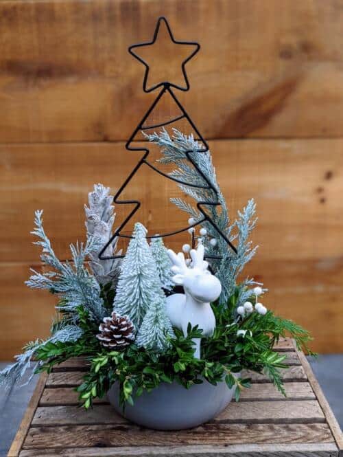 A grey cermaic container filledw ith holiday greenery adorned with white bottle brush trees, a white ceramic reindeer and large black wire tree with a star on top.