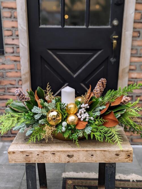 a large oblong gold container filled with winter greenery and gold ornaments adorned with a large white pillar candle