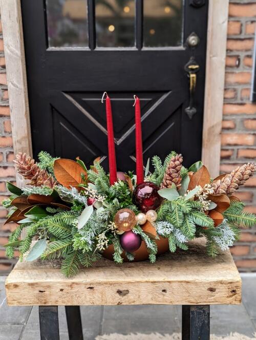 a large oval gold ceramic overflowing with winter greenery adorned with red and burgundy ornaments and red tapered candles