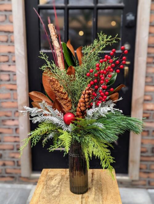 A dark glass vase filled with winter greenery, red berries and a red bell.