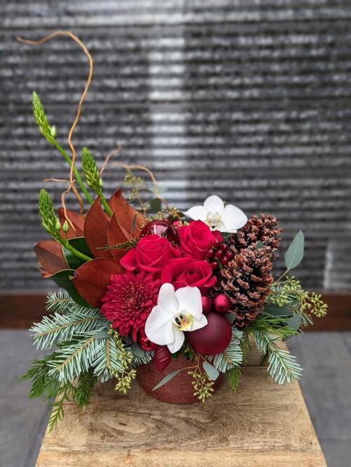 A red metalllic container filled with red florals and christmas greenery. featuring red roses, star of bethlehem, magnolia and red ornaments.