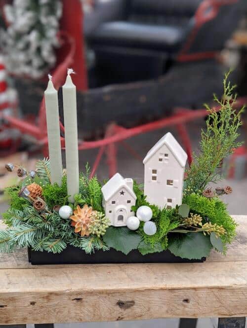 A black shallow dish overflowing with winter greenery adorned with 2 white ceramic houses and light green candles.