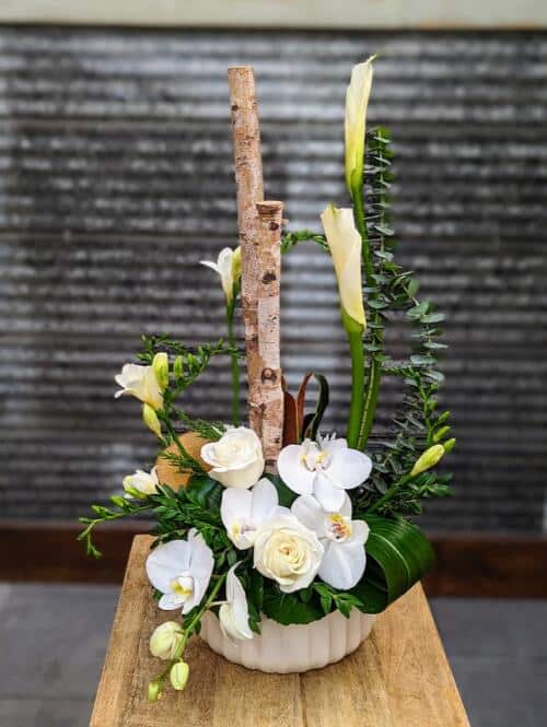 a large white ceramic container filled with white and green blooms featuring calla lilies, orchids, freesia and birch poles